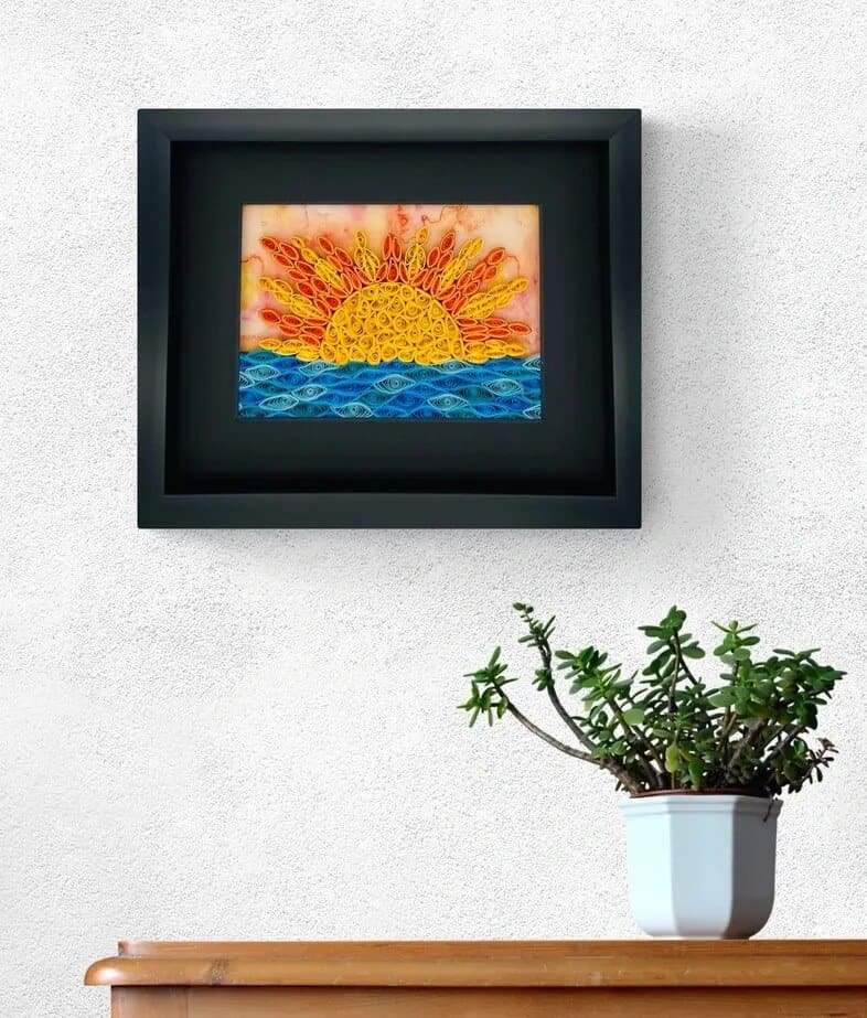 Framed artwork of a sun rising over blue waves, displayed on a white wall above a potted plant.