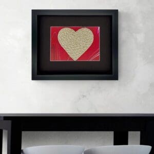 Framed heart-shaped art with handwritten text on a red background above a black desk.